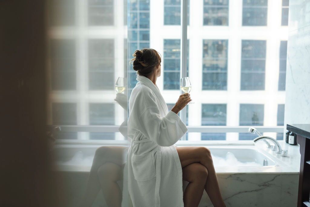 A person in a white bathrobe sitting by a window with a view of buildings, holding a glass of white wine.