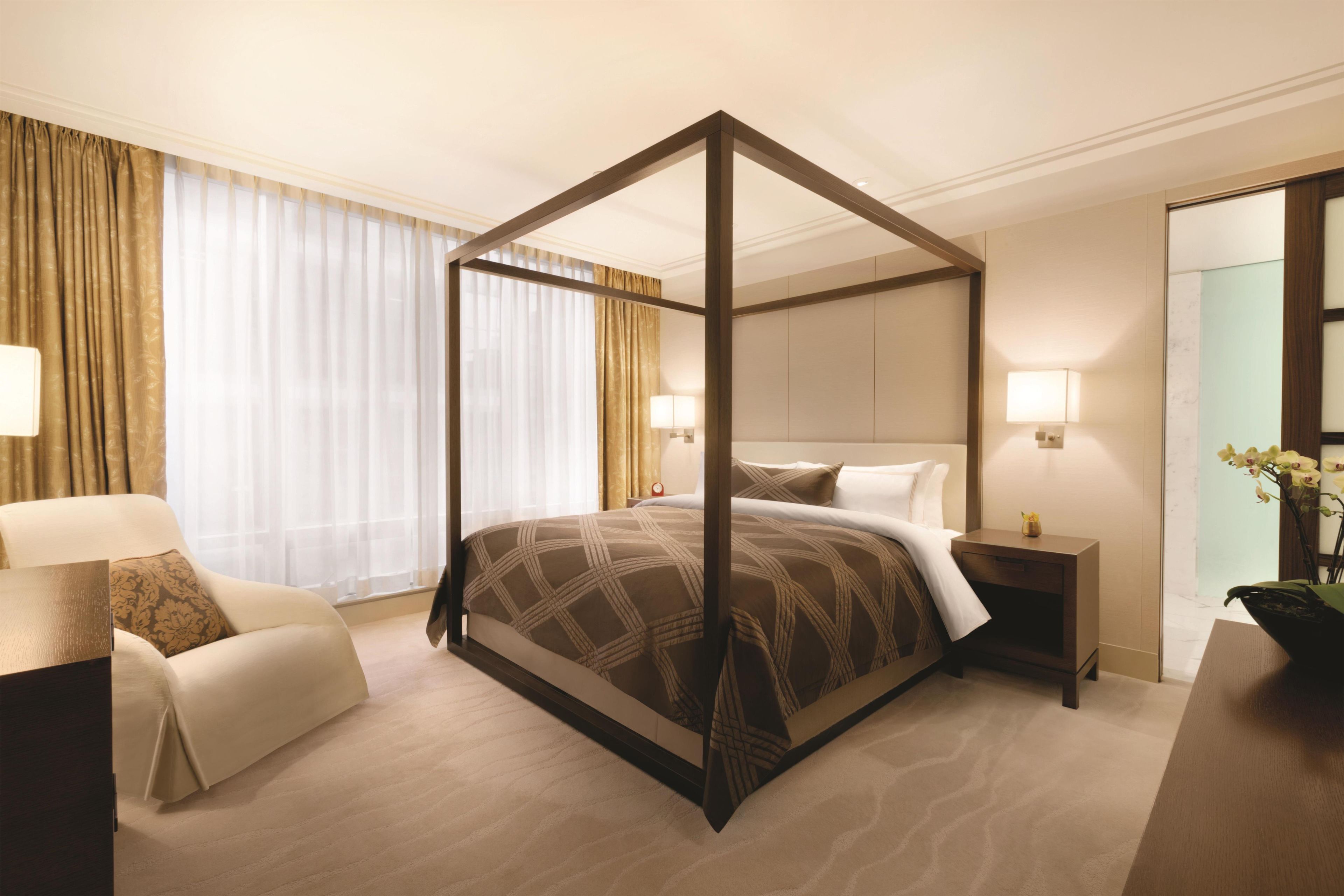A four-poster bed with a checkered bedspread, a bedside table with a lamp, a chair with a cushion, and a large mirror on the wall.
