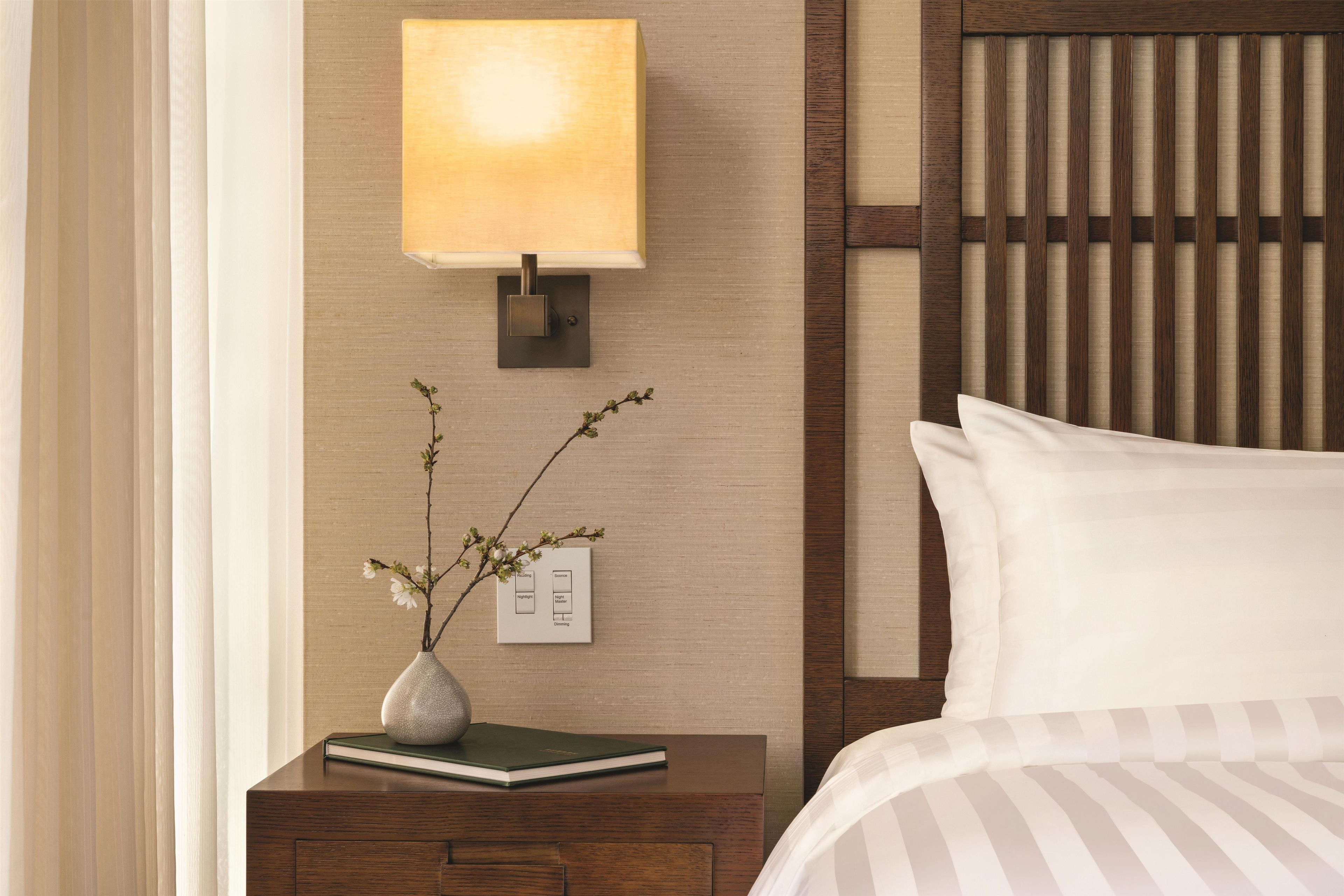 A neatly arranged hotel room with a bedside table, a lamp, a vase with flowers, and a bed with white bedding.