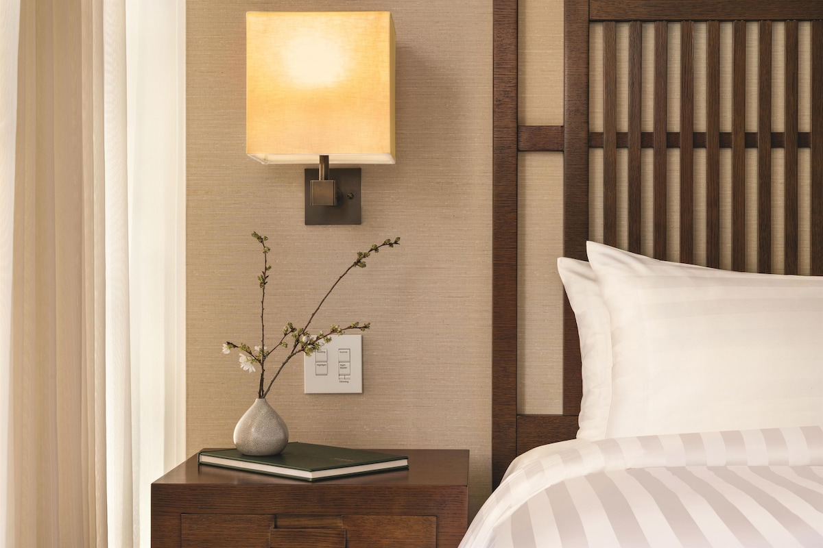 Une chambre d’hôtel bien rangée, avec une table de chevet, une lampe, un vase de fleurs et un lit recouvert de draps blancs.