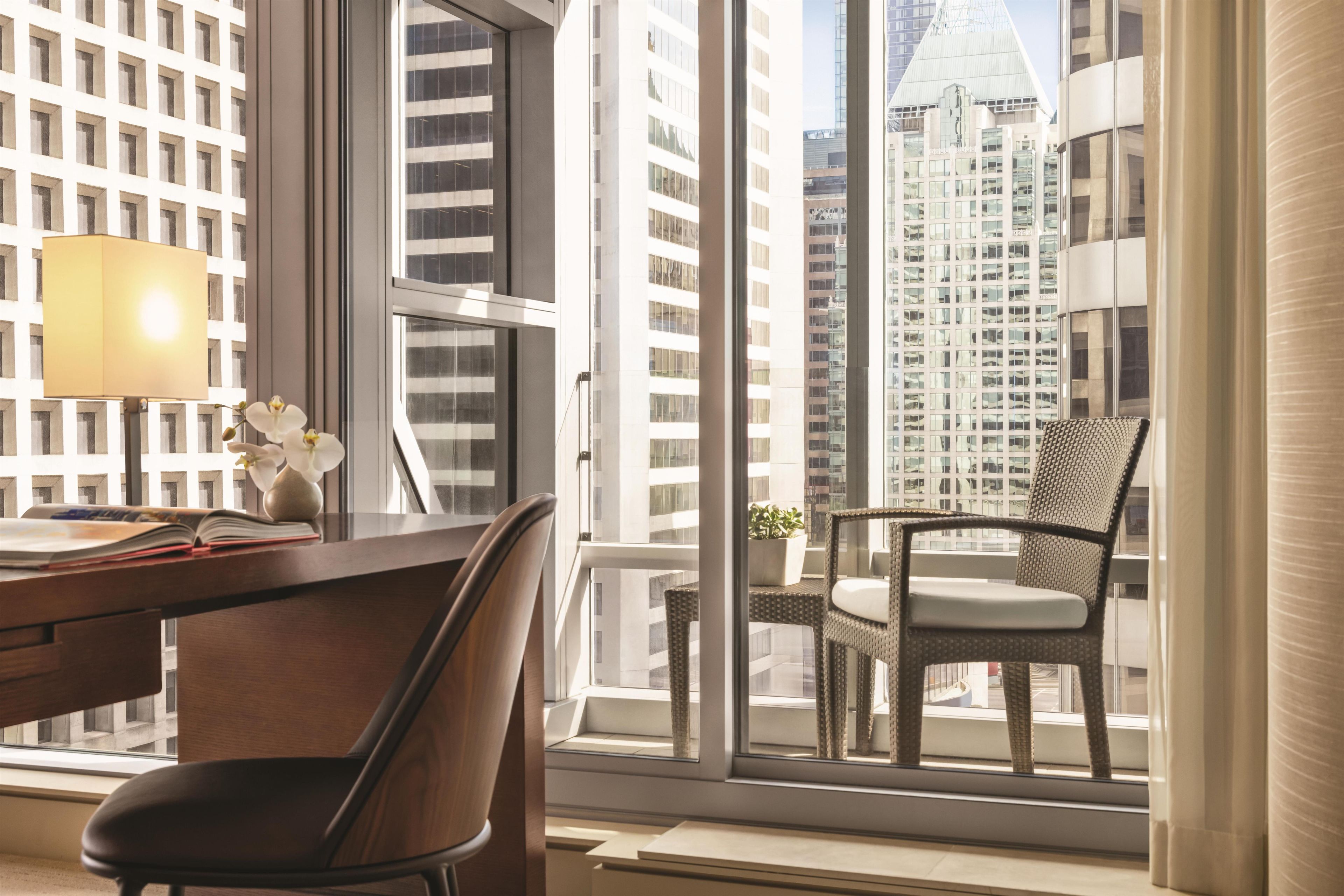 Habitación de hotel moderna con un ventanal grande que ofrece vistas a la ciudad, un escritorio con lámpara, una silla y una pequeña mesa con una planta.