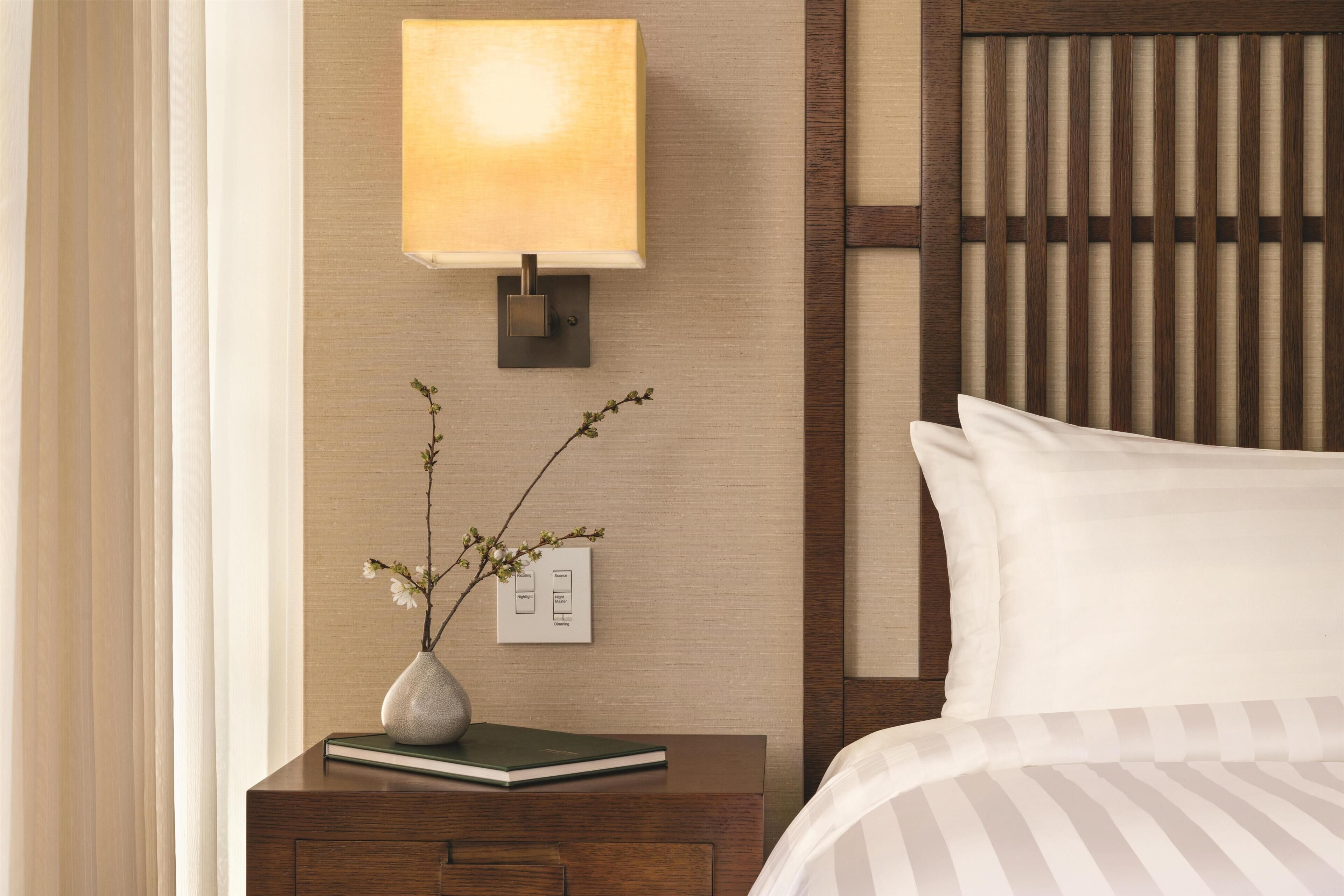 Una habitación de hotel ordenada con una mesita de noche, una lámpara, un florero con flores y una cama con ropa blanca.