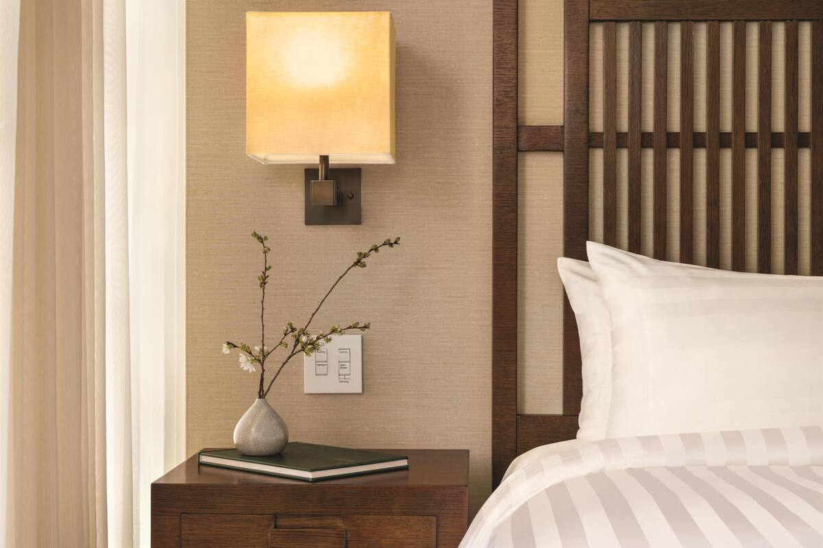 Une chambre d’hôtel bien rangée, avec une table de chevet, une lampe, un vase de fleurs et un lit recouvert de draps blancs.