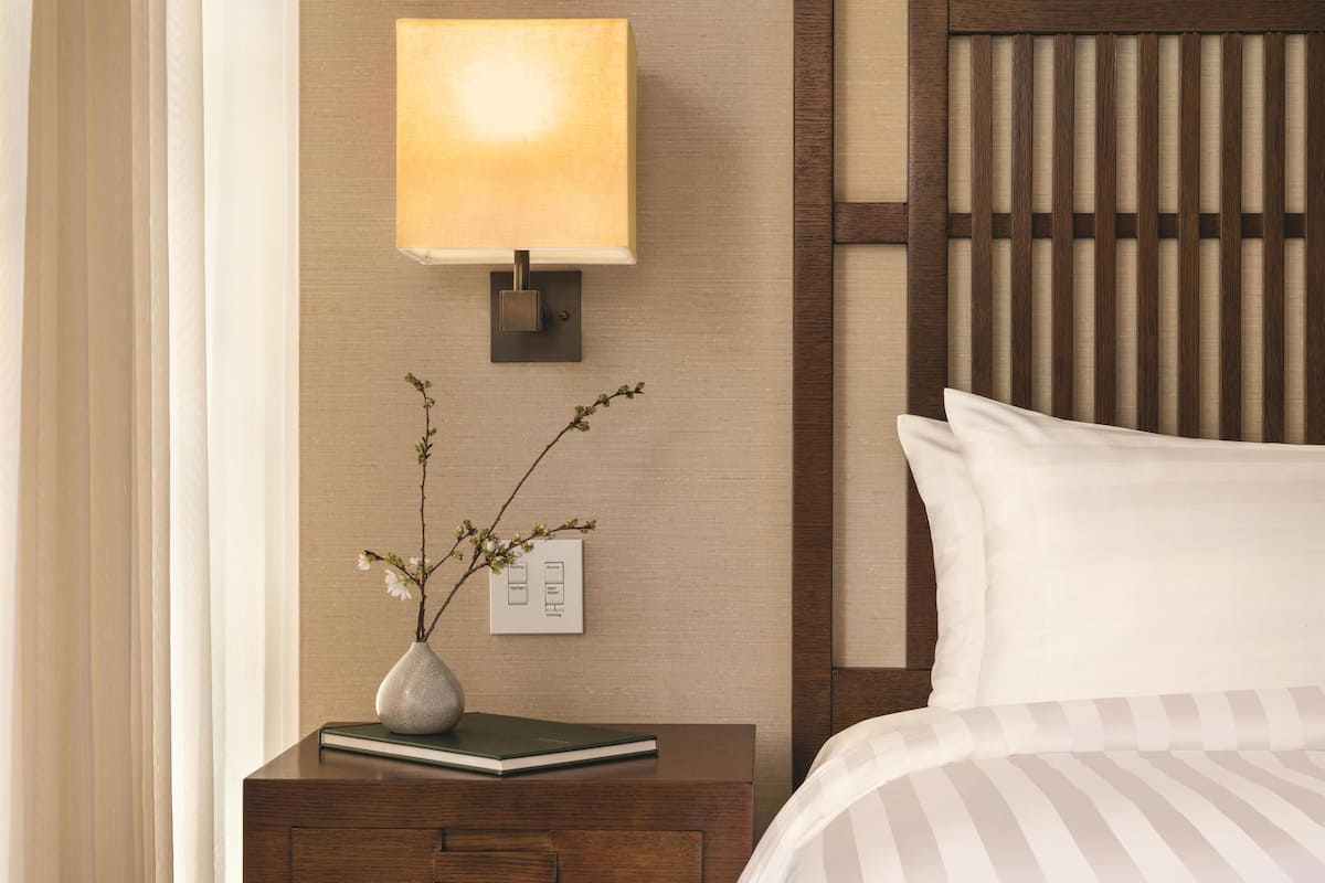 Ein ordentlich eingerichtetes Hotelzimmer mit Nachttisch, Lampe, Blumenvase und einem Bett mit weißer Bettwäsche.