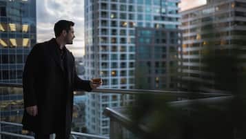 A man in a dark coat holding a drink on a balcony with a cityscape view.