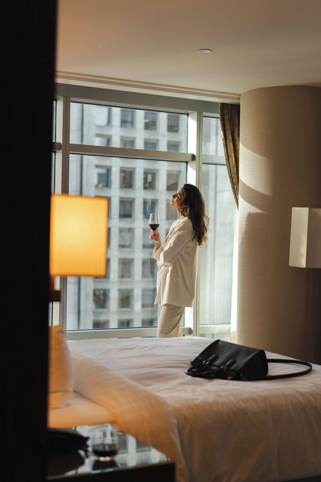 A hotel room with a large window, a bed, a bedside table with a glass of wine, and a black bag on the bed.