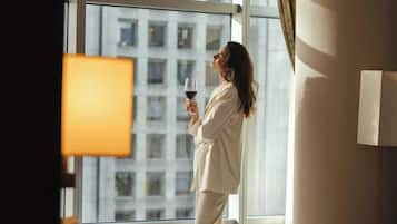 A hotel room with a large window, a bed, a bedside table with a glass of wine, and a black bag on the bed.