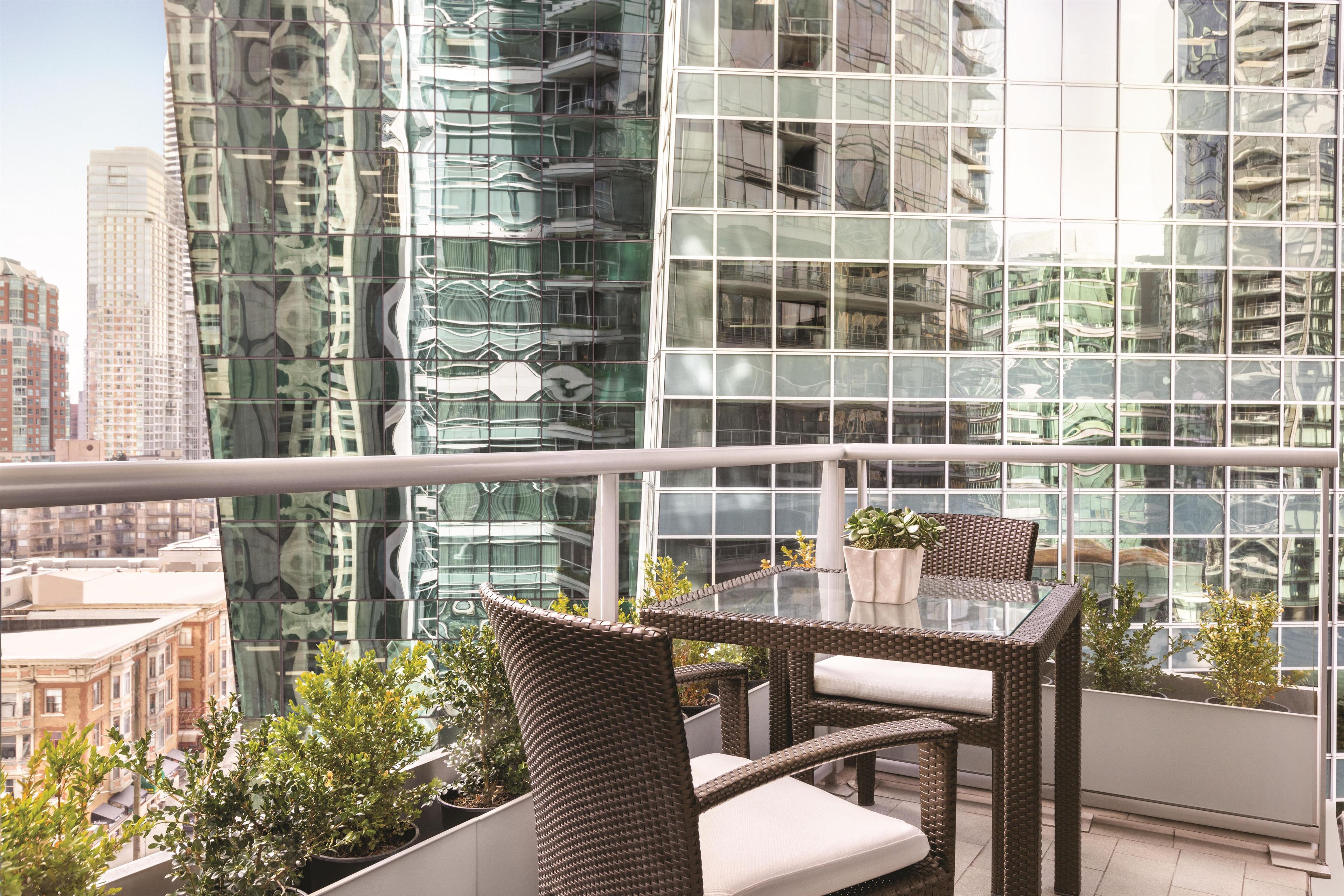 A balcony with wicker furniture and potted plants, overlooking a cityscape with modern buildings.