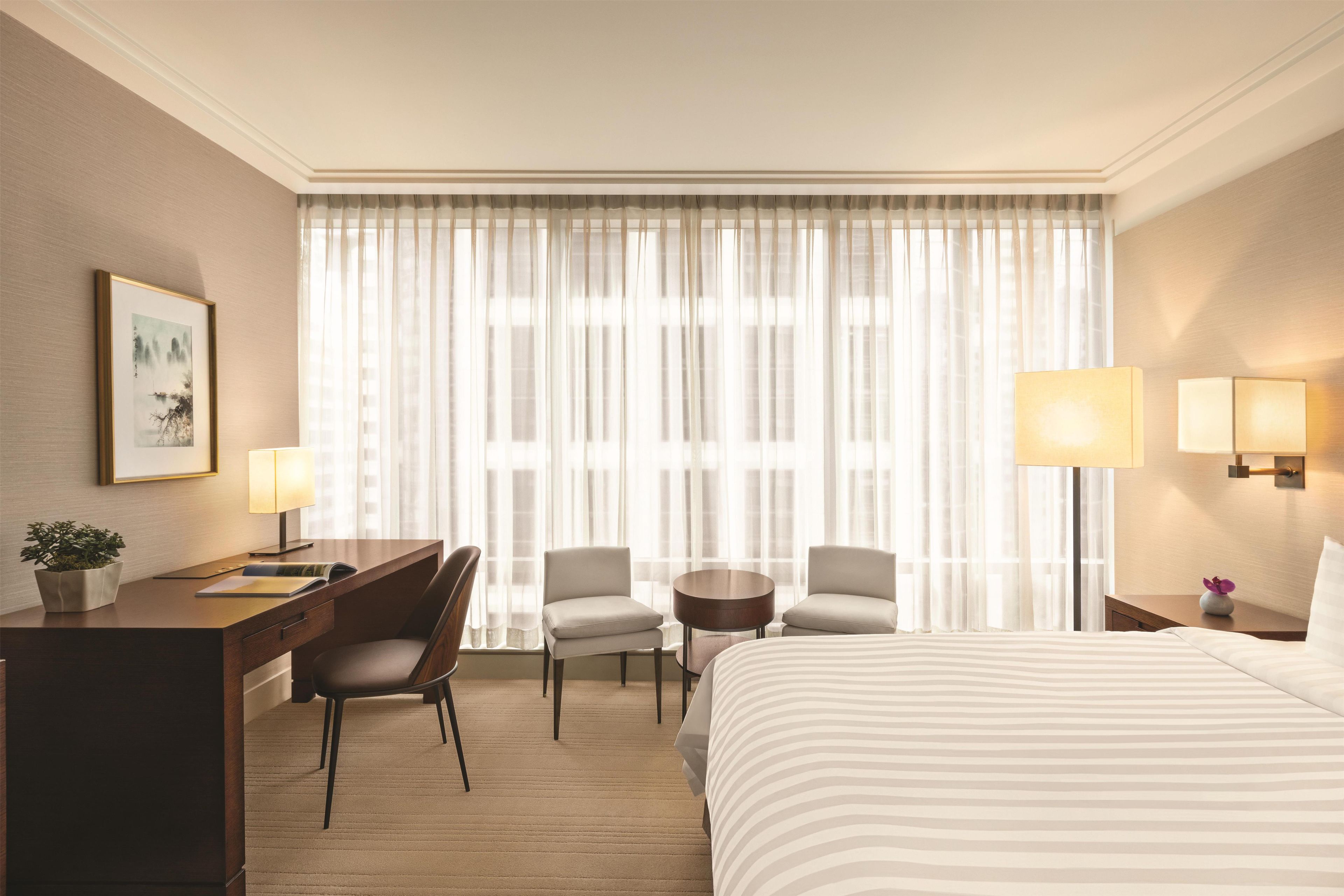 A hotel room with a bed, two chairs, a desk, a small table, a lamp, and a window with sheer curtains.