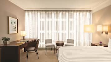 A hotel room with a bed, two chairs, a desk, a small table, a lamp, and a window with sheer curtains.