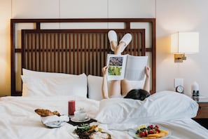 Una persona acostada en la cama leyendo un libro, con una bandeja de desayuno a su lado.