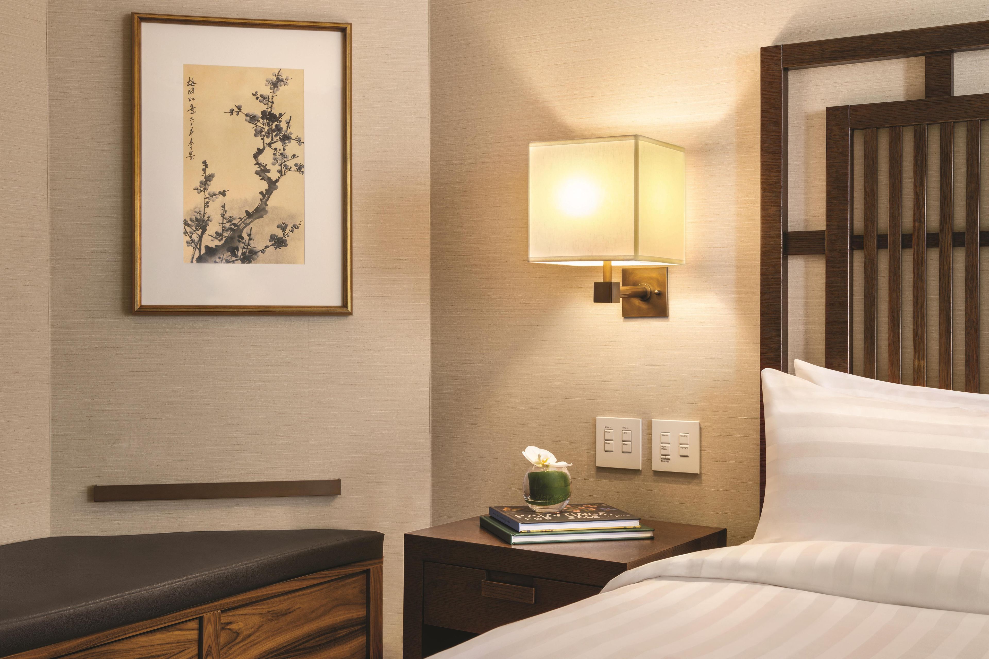 A hotel room with a bed, bedside table, lamp, framed artwork, and a small vase with a flower.