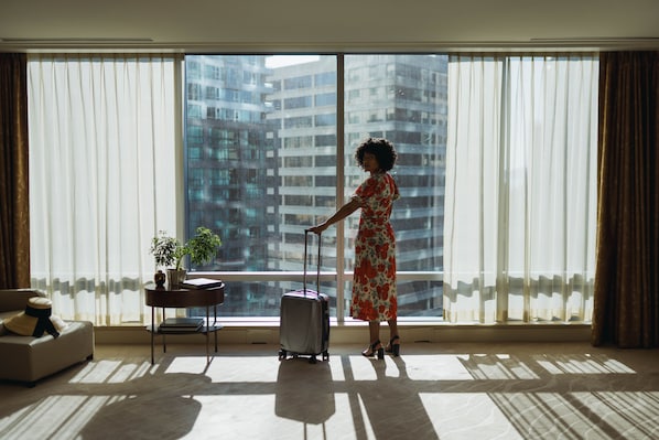 A room with a large window, a suitcase, a sofa, and a small table with a plant.