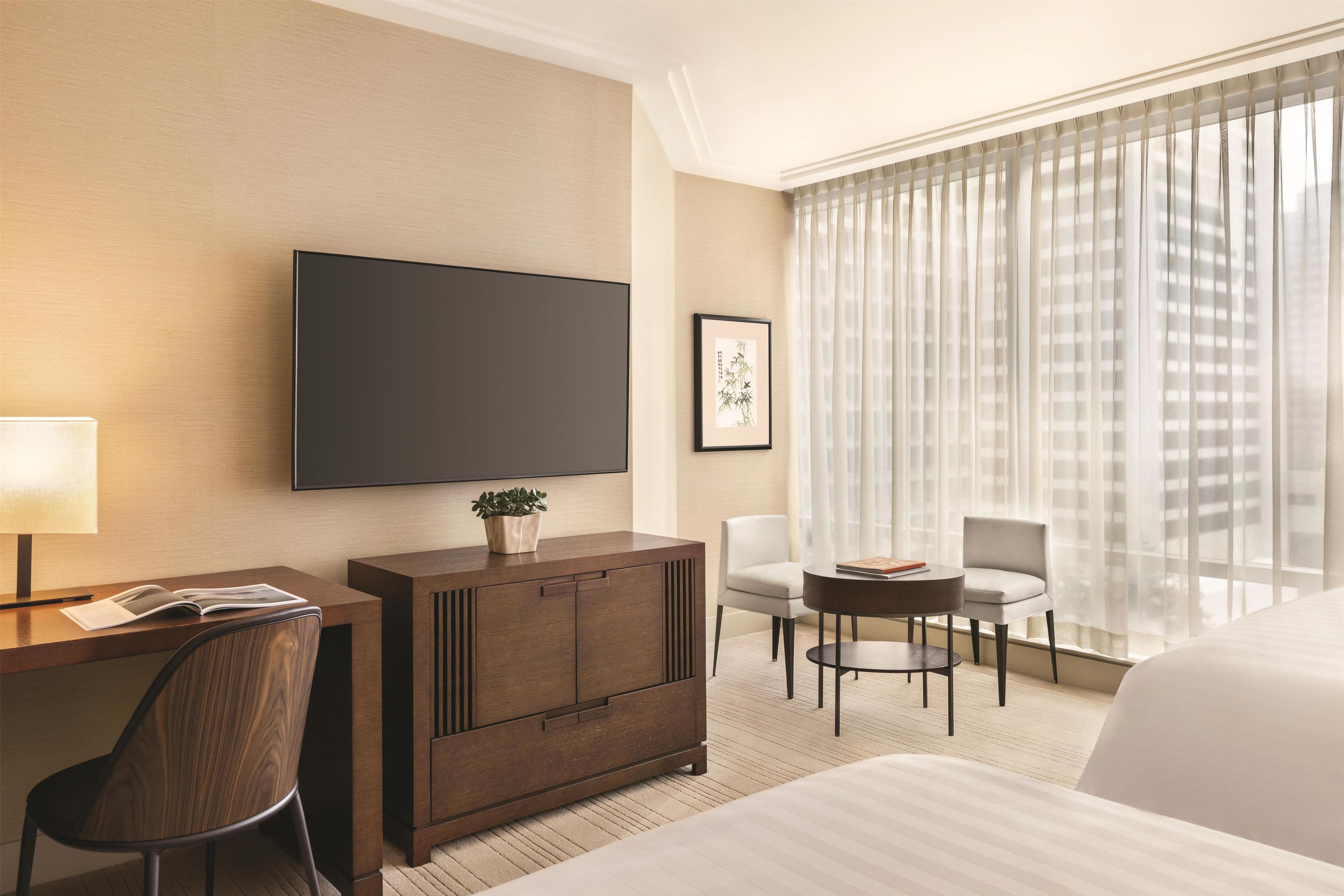 A hotel room with a large bed, a desk with a chair, a TV, a small table with chairs, and a window with sheer curtains.