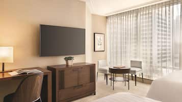 A hotel room with a large bed, a desk with a chair, a TV, a small table with chairs, and a window with sheer curtains.