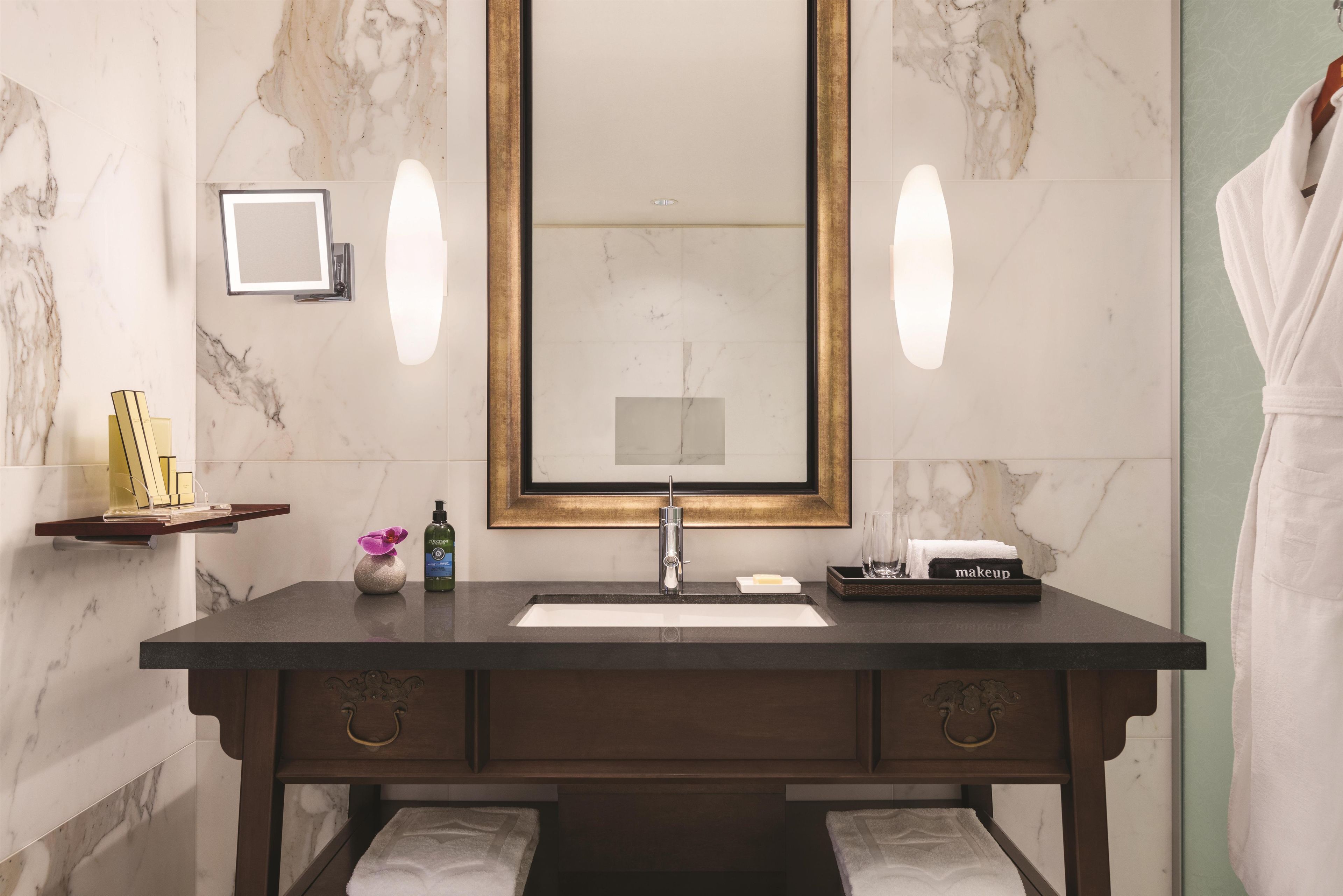 A bathroom with a marble wall, a dark wooden vanity, a large mirror, and a robe hanging to the side.
