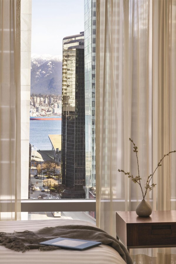 A bedroom with a large window, a bed, a bedside table with a vase, and a view of a cityscape and mountains.