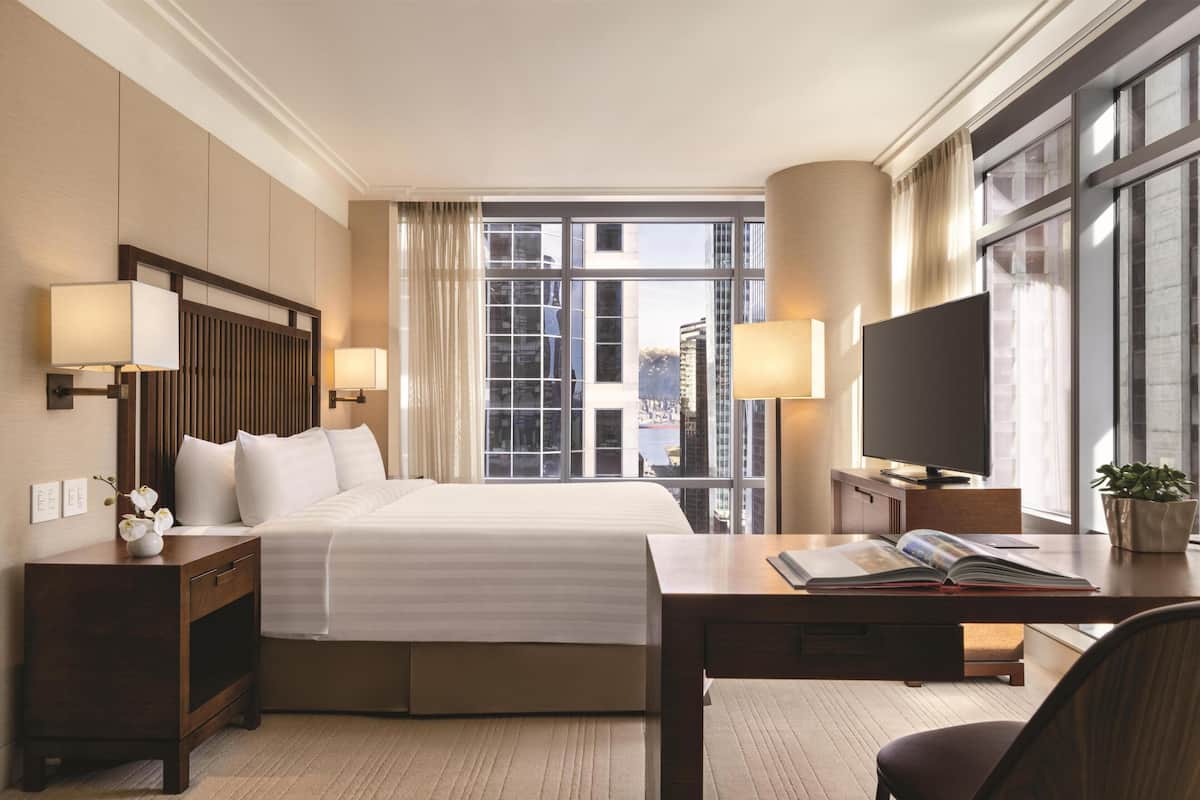 Une chambre d’hôtel avec un grand lit, deux tables de chevet avec des lampes, une télévision, un bureau avec un livre, et une vue sur la ville grâce à de grandes fenêtres.