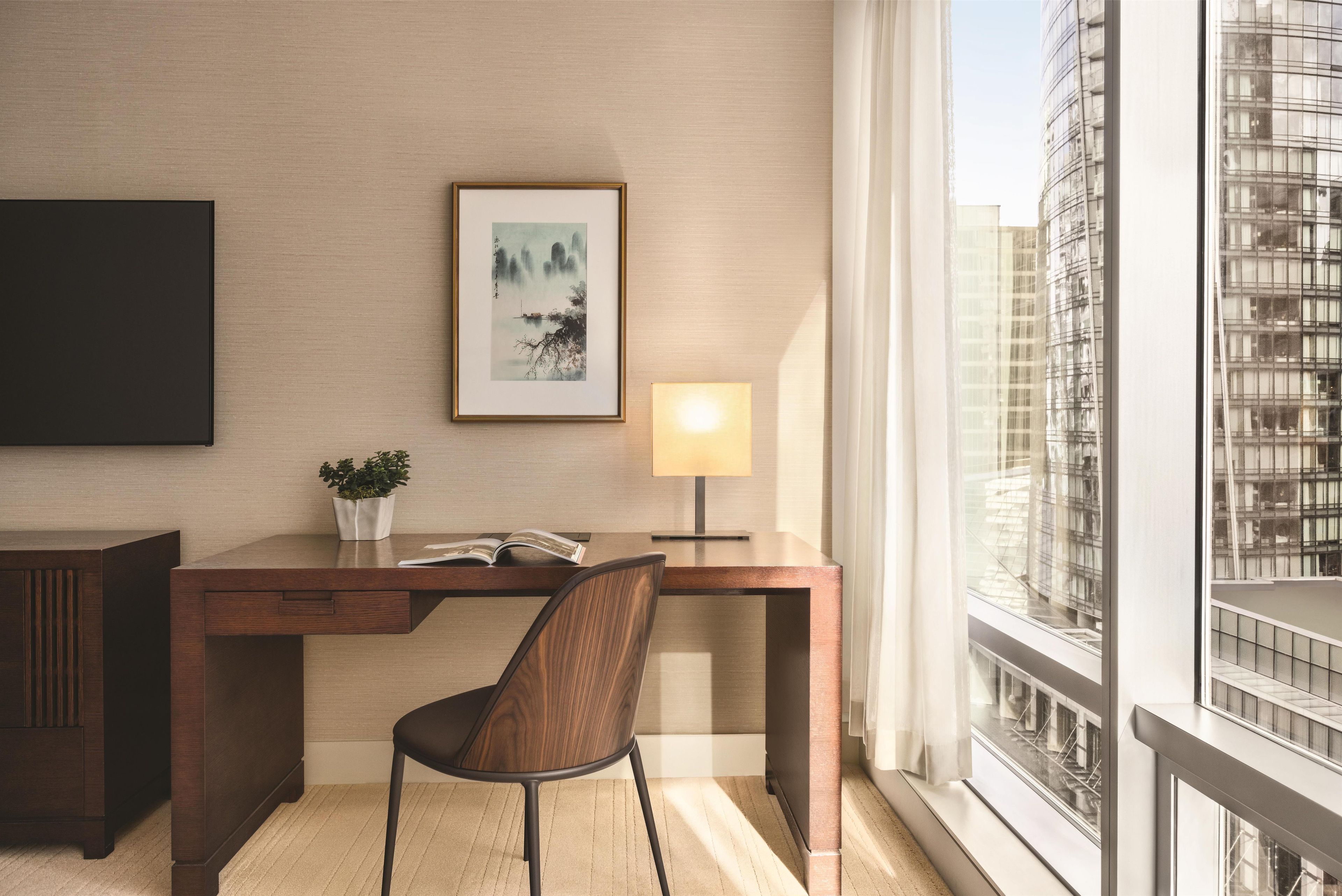 Una habitación de hotel moderna con un escritorio de madera, una silla, una obra de arte enmarcada, una lámpara de noche y vistas a un paisaje urbano a través de amplios ventanales.