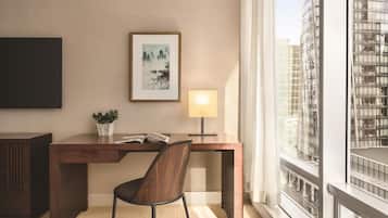 Una habitación de hotel moderna con un escritorio de madera, una silla, una obra de arte enmarcada, una lámpara de noche y vistas a un paisaje urbano a través de amplios ventanales.