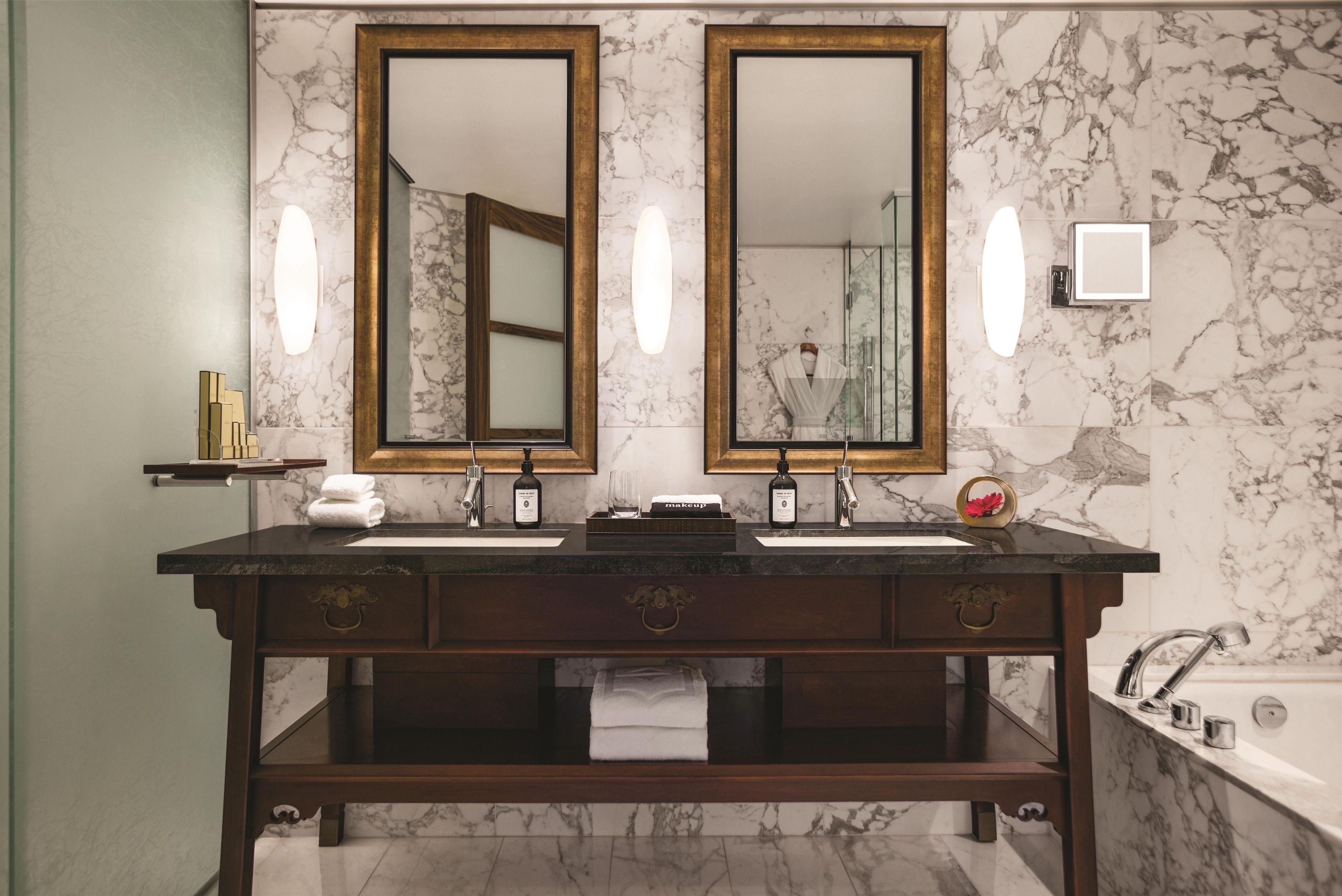 A bathroom with two mirrors, marble walls, a dark wooden vanity, a bathtub, and a towel rack.