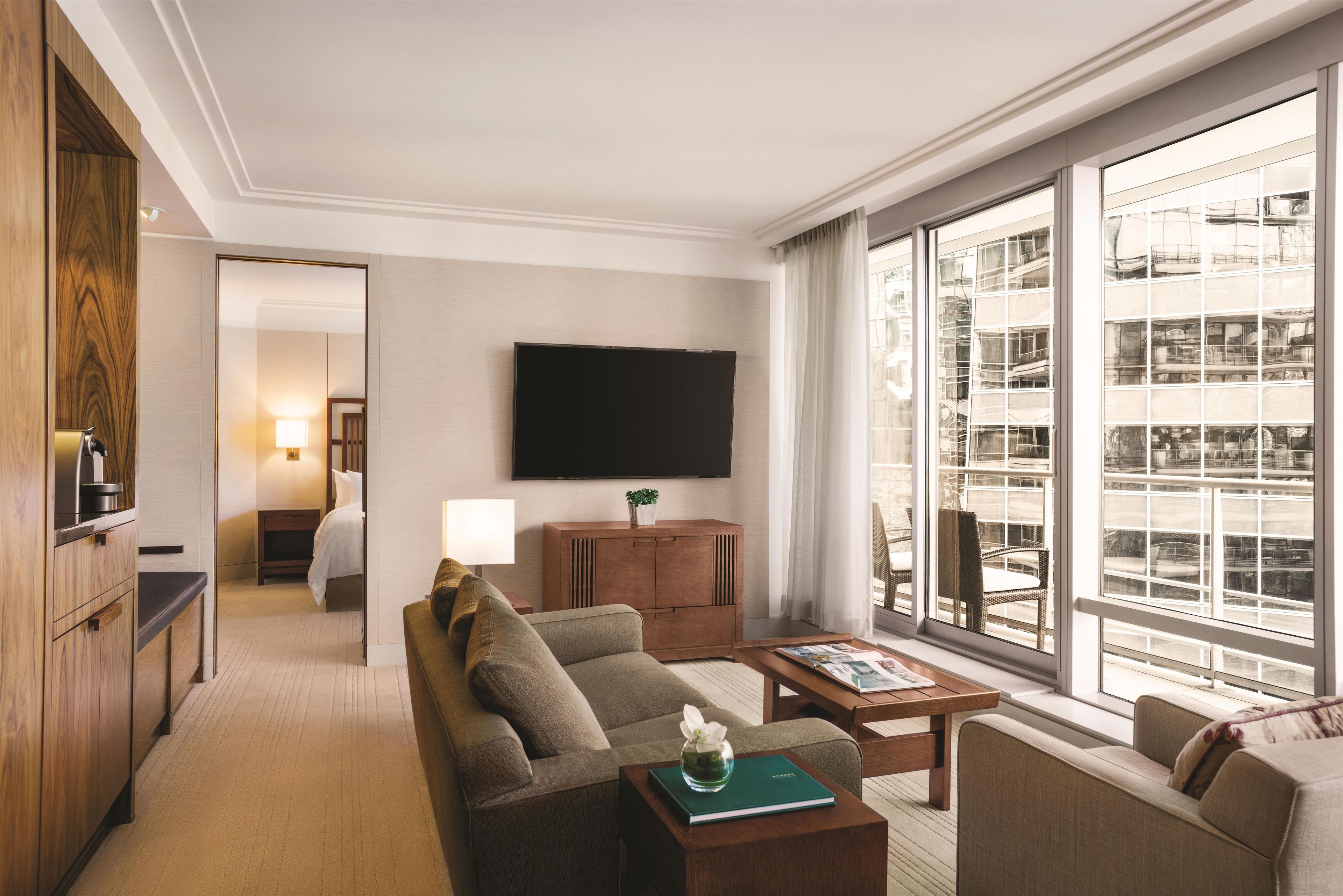 A hotel room with a sofa, coffee table, and a TV mounted on the wall. There is a window with a view of buildings.