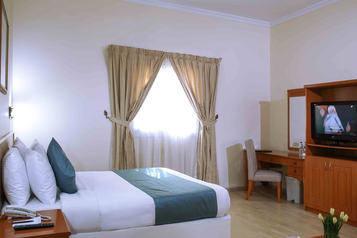 A hotel room with a bed, a desk, a chair, a television, and a small table with a phone and remote control.