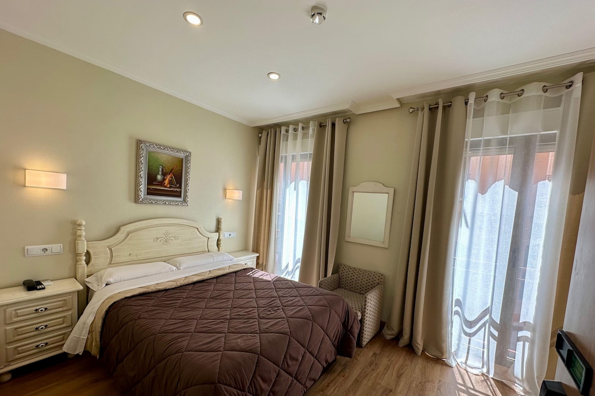 A hotel room with a large bed, bedside tables, a nightstand, a chair, a painting on the wall, and a window with curtains.