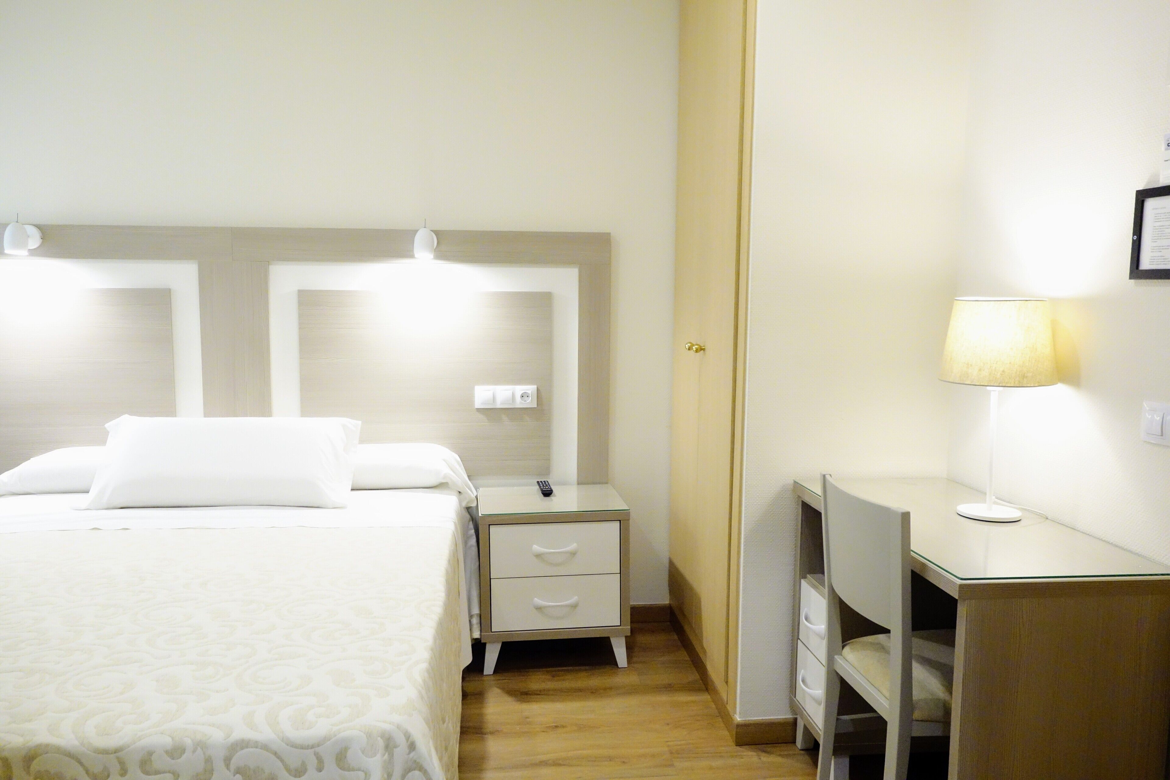 A hotel room with a bed, bedside tables, a desk with a lamp, and a door.