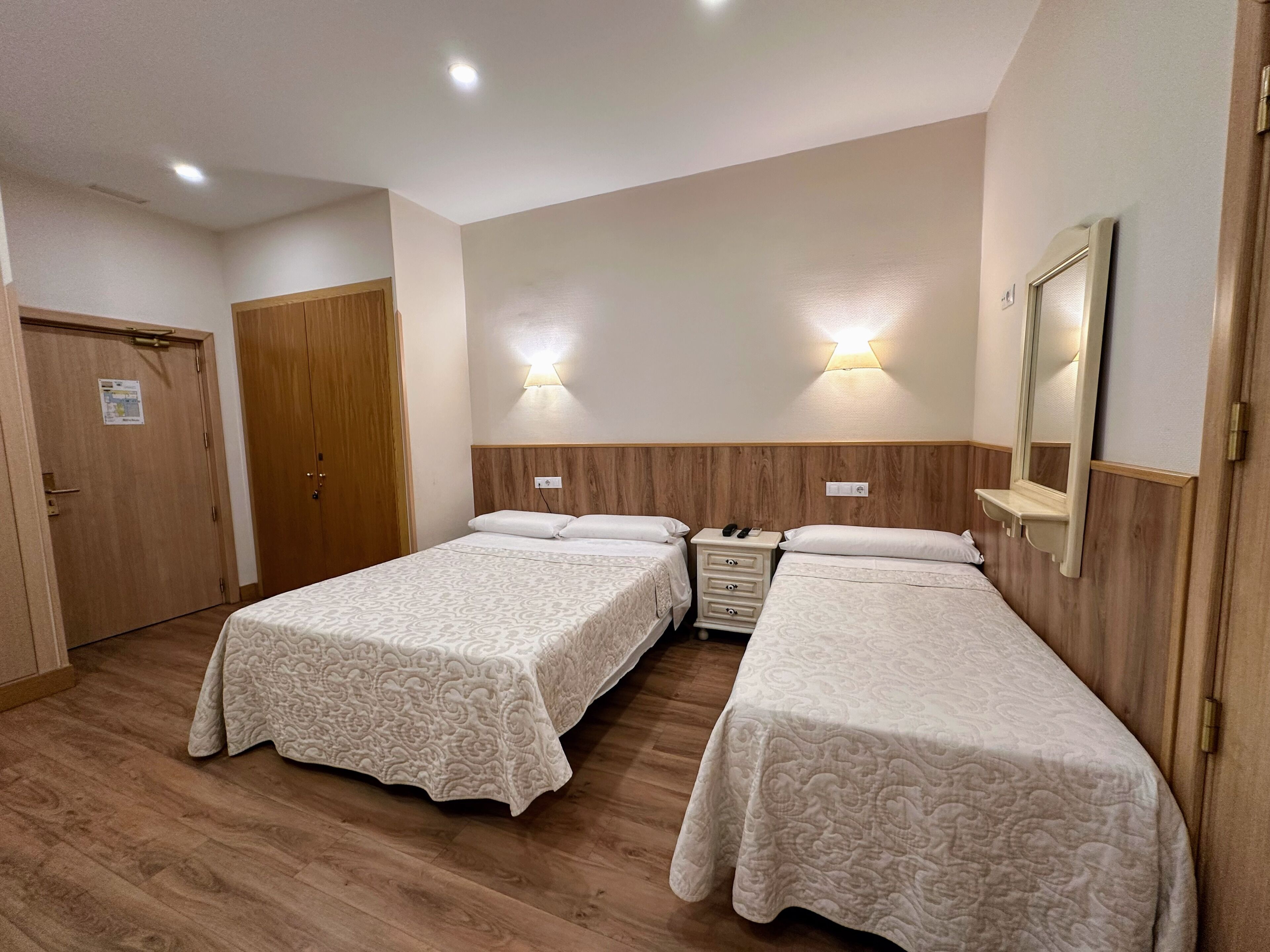 A hotel room with two single beds, wooden headboards, a wooden wardrobe, and a mirror.