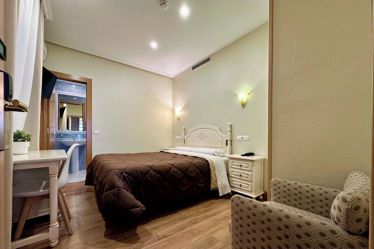 A bedroom with a bed, bedside table, chair, and a desk with a chair.
