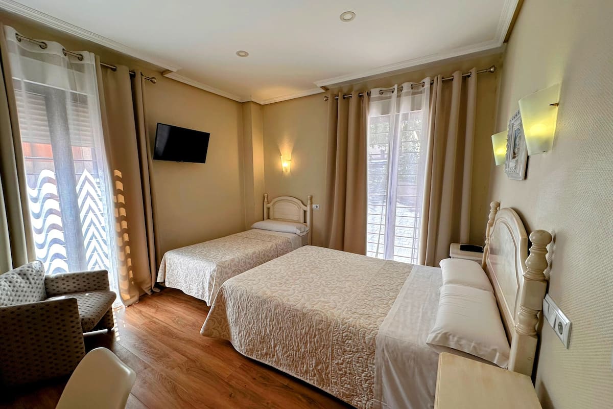A hotel room with two single beds, a TV mounted on the wall, a chair, and a window with curtains.