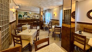 A restaurant with multiple tables and chairs, a counter with a person serving, and a view of the kitchen through a glass wall. - Hostal Toledo (Toledo)