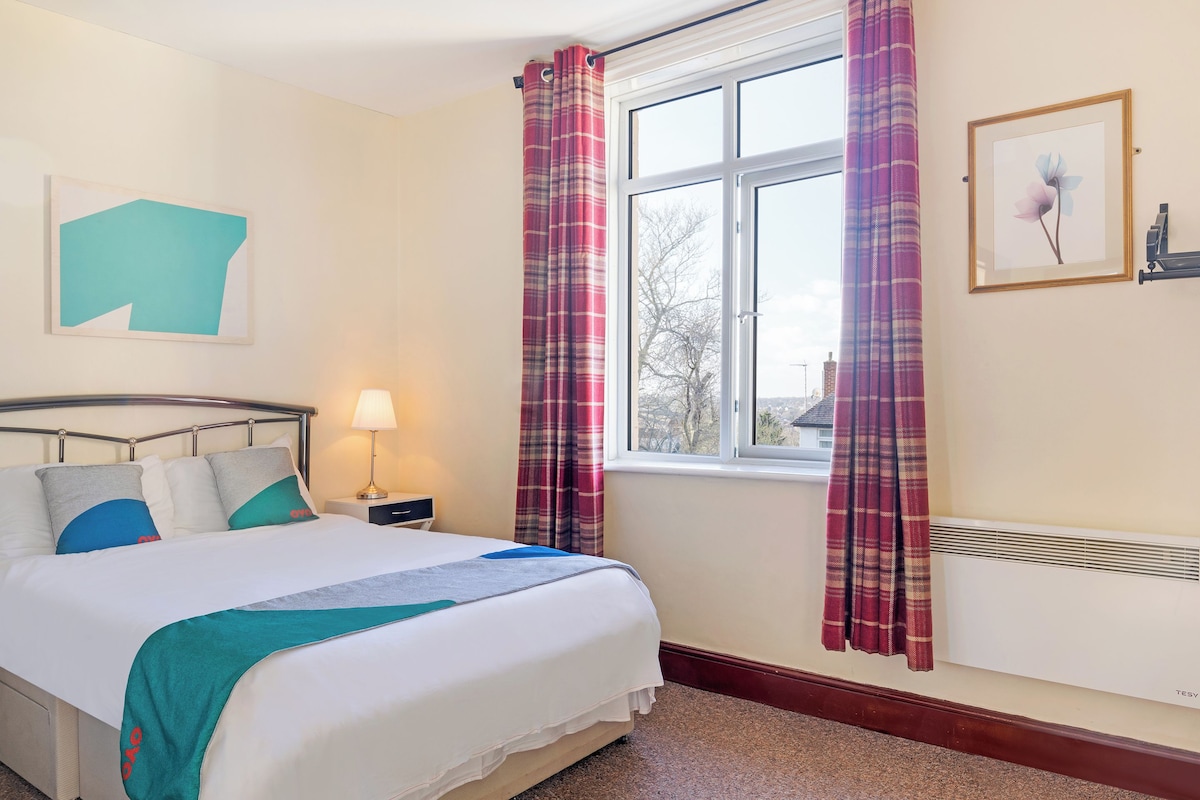 A bedroom with a bed, a nightstand, a lamp, a window with curtains, a radiator, and a framed picture on the wall.
