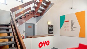 A hotel reception area with a wooden staircase, a reception desk with the OYO logo, a multilingual welcome sign, and a red chair. - OYO The Green Man Pub and Hotel (Wembley)