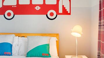 A bed with a wooden headboard, white bedding, and colorful pillows. A red wall decal of a bus is above the bed. A white bedside table with a lamp is to the right.
