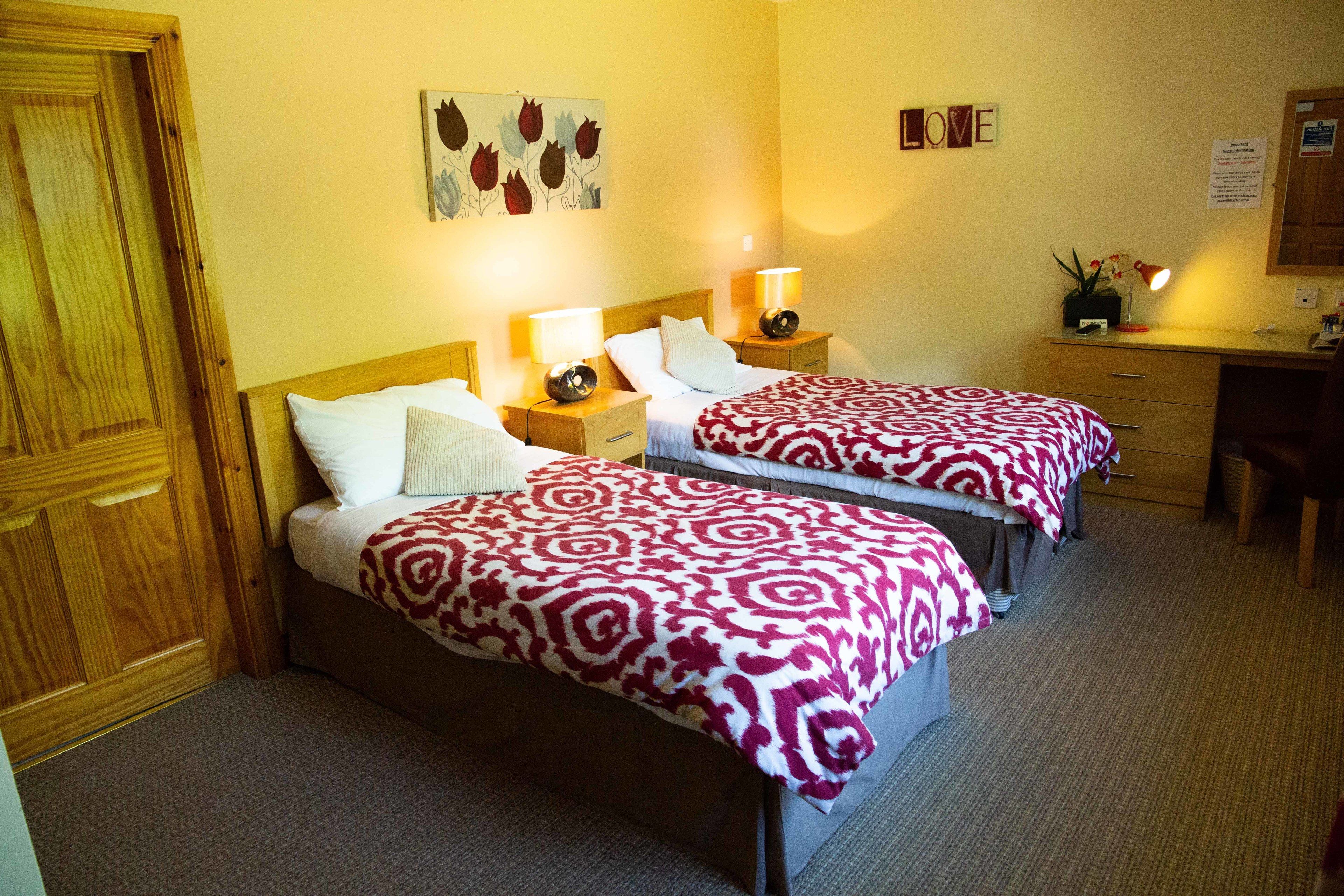 A hotel room with two beds, wooden headboards, bedside tables with lamps, a desk, and a chair.