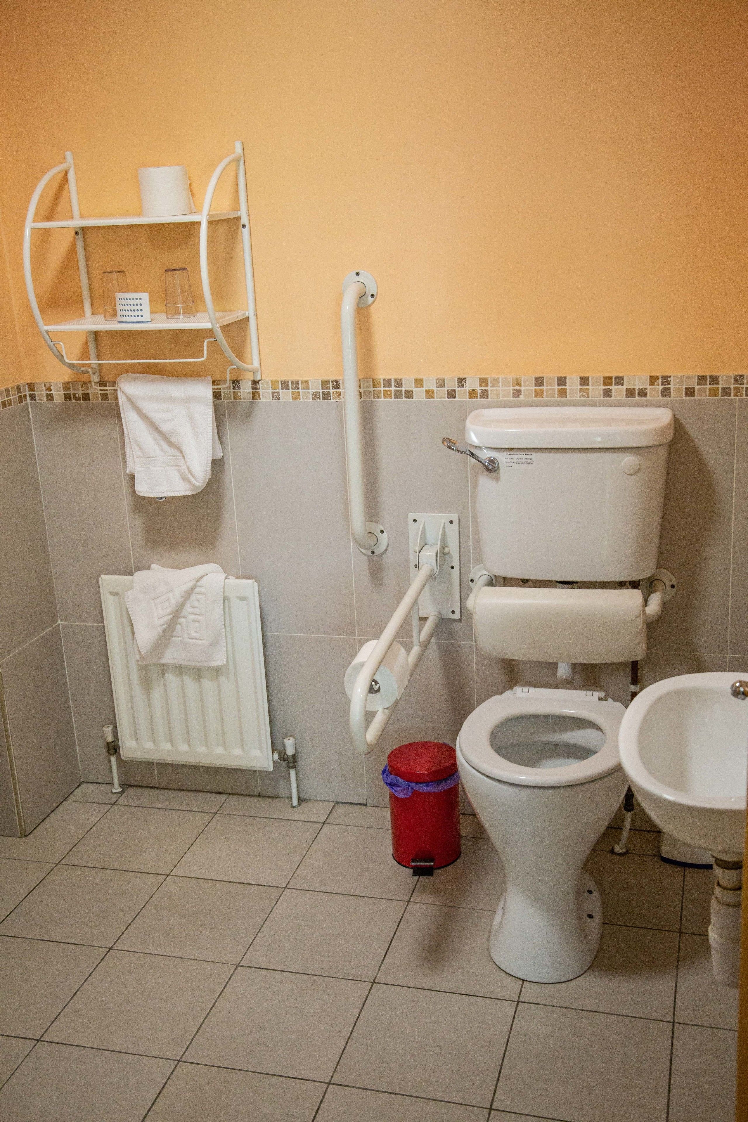 A bathroom with a toilet, bidet, and sink. There are shelves with toiletries and a red trash bin.