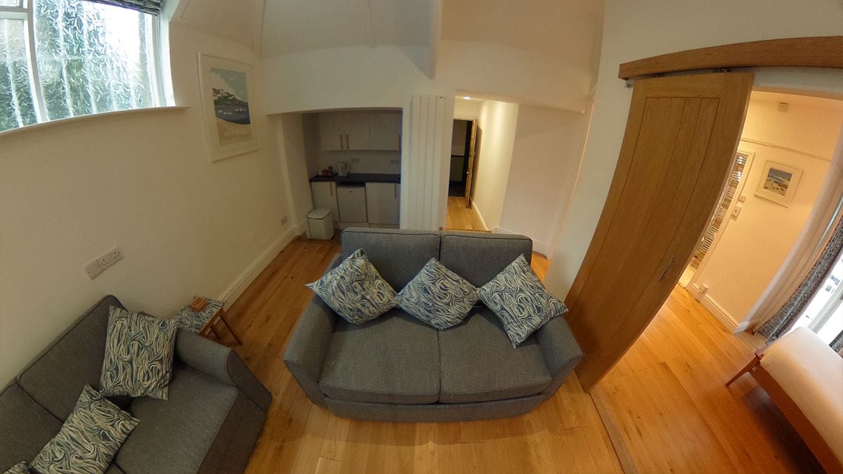 Ein Wohnzimmer mit einer grauen Sofagarnitur, Holzboden und einer Küche im Hintergrund.