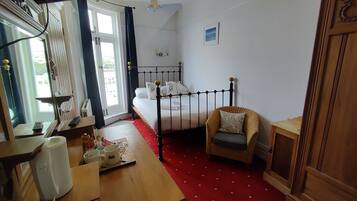 Un dormitorio con cama, una silla, una mesa con un tapete rojo y una ventana con cortinas.