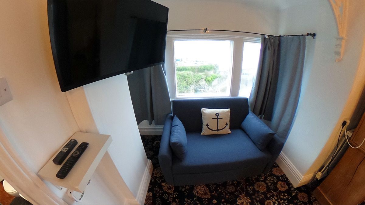 A small room with a blue sofa, a TV mounted on the wall, a window with curtains, and a remote control on a small table.