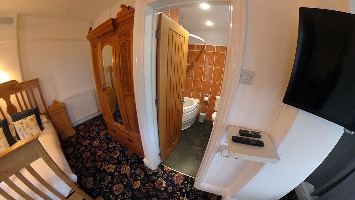 Ein Badezimmer mit Toilette, Waschbecken und Dusche. Es gibt einen Holzschrank und einen an der Wand befestigten Fernseher.