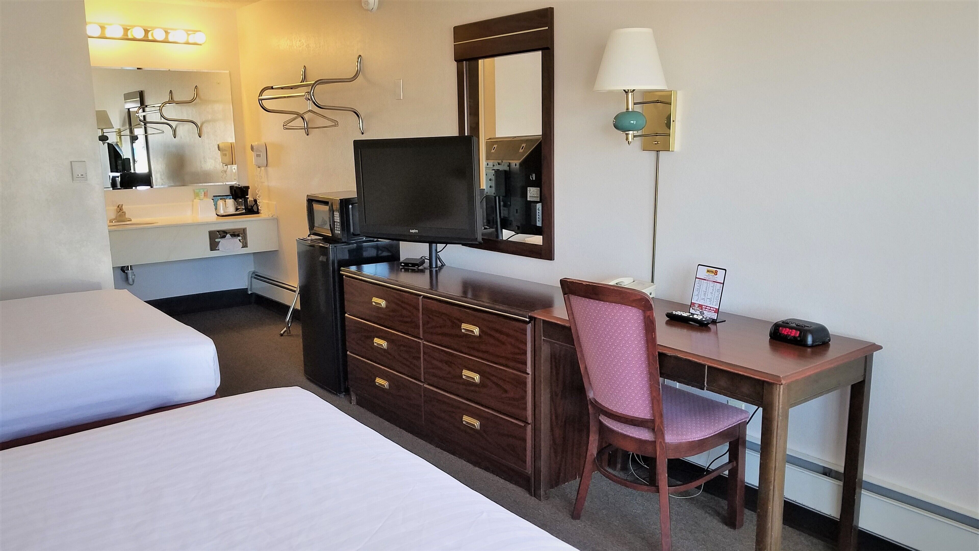 Une chambre d’hôtel avec deux lits, un bureau avec une chaise, une télévision, un miroir, une lampe et un mini-bar.