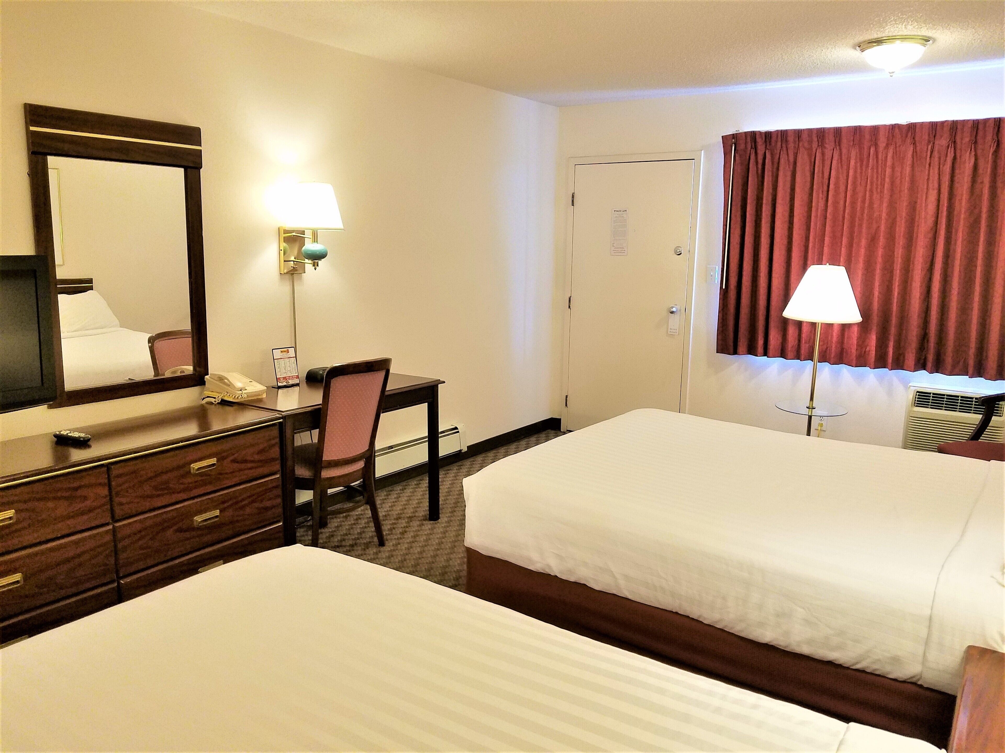 Quarto de hotel com duas camas, uma mesa com cadeira, uma luminária de mesa, um telefone, um espelho e uma janela com cortinas.