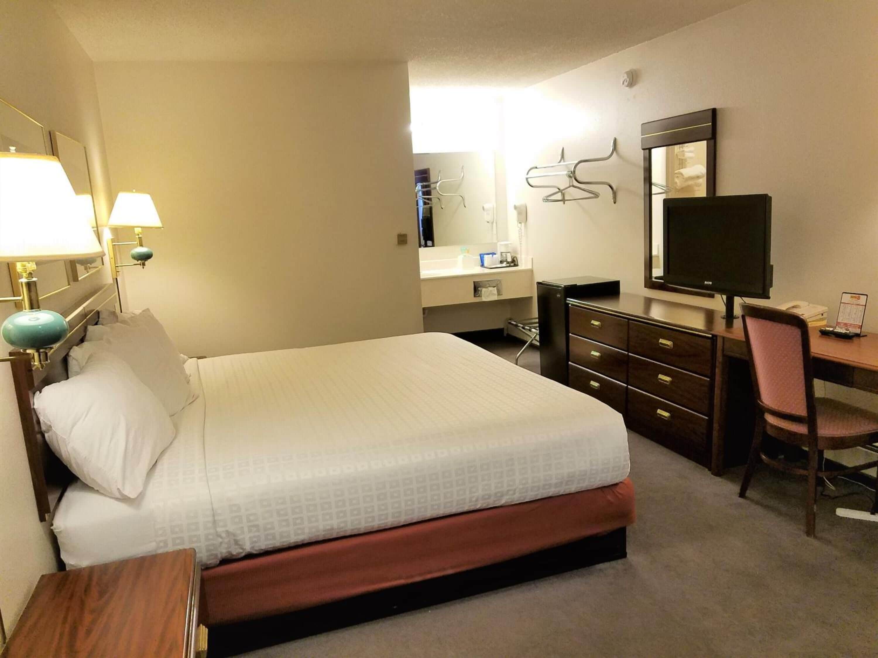 A hotel room with a bed, a desk with a computer, a chair, a dresser, a mirror, and a lamp.