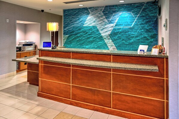 Un taulell de recepció d'hotel amb una pantalla digital, una làmpada i un panell d'anuncis.