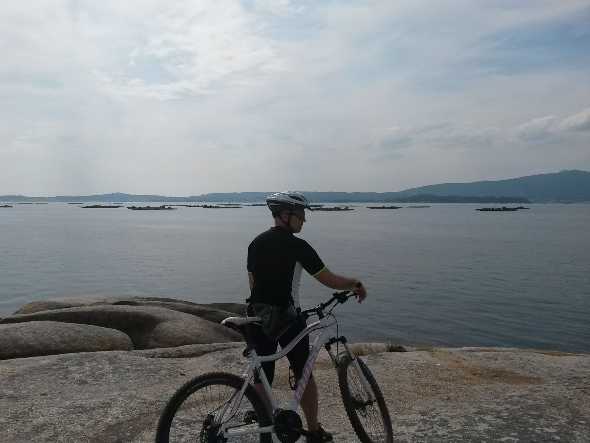 A person standing on a rocky shore with a bicycle, overlooking a calm body of water with several boats and distant hills.