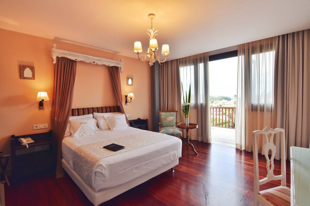 A hotel room with a large bed, a chandelier, a balcony with a view, and a chair.