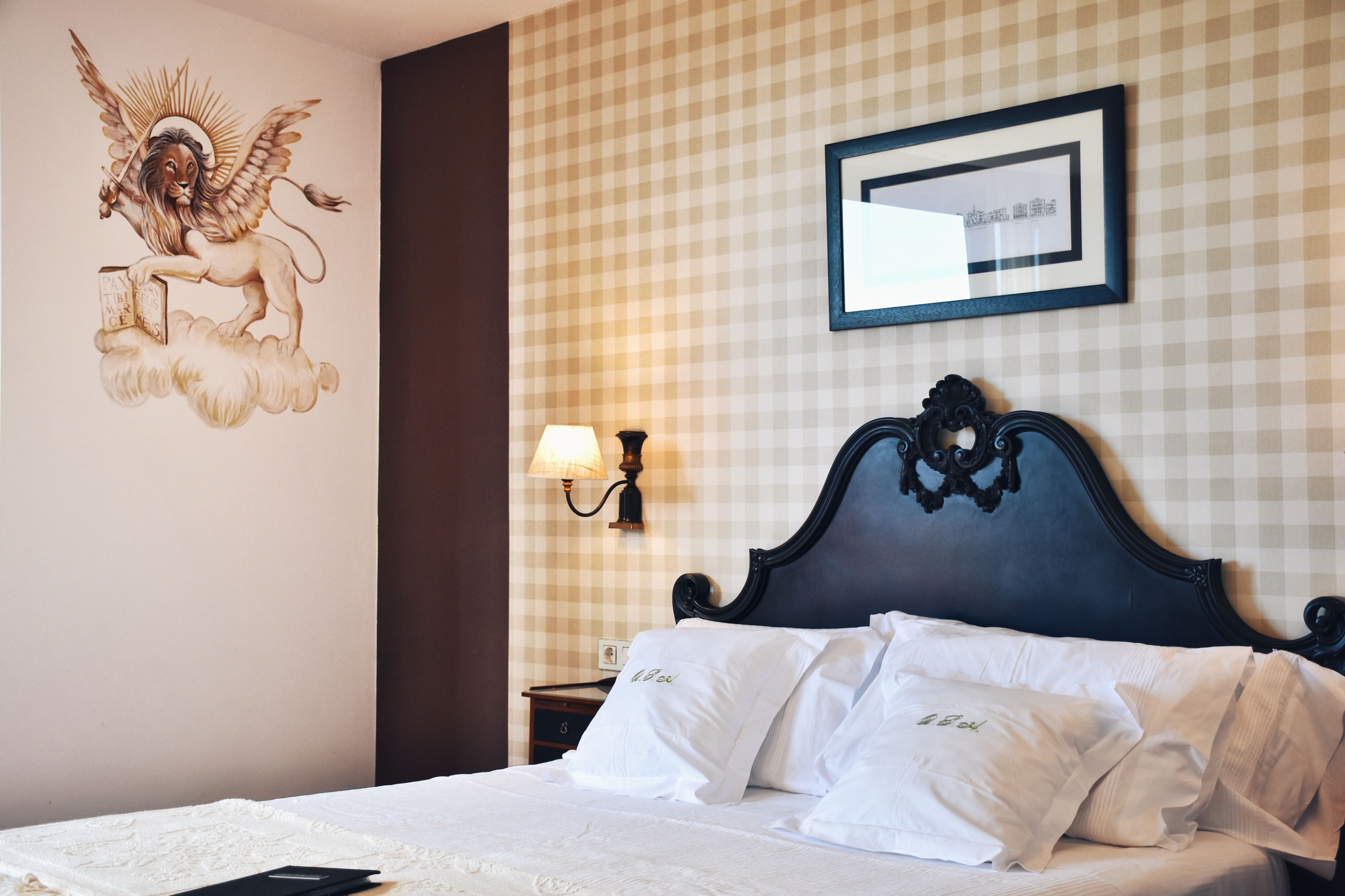 Hotelkamer met een geruit wandpaneel, een decoratieve engelmuur, een ingelijst beeld en een klassiek bed met witte lakens, plus een nachtkastje met een lamp.