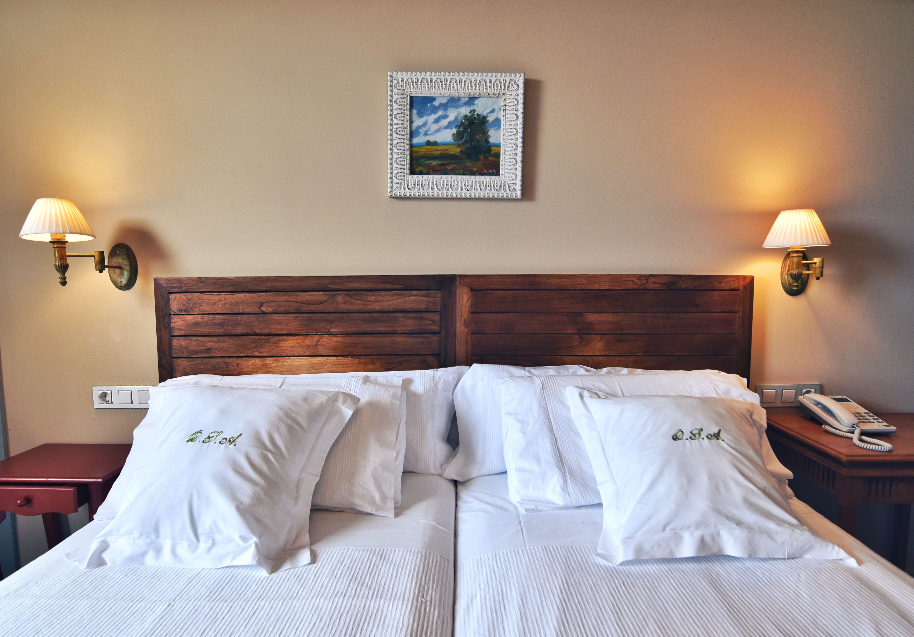 Una cama doble con ropa de cama y almohadas blancas, un cabecero de madera, lámparas de noche, un cuadro en la pared y una mesita de noche con un teléfono.